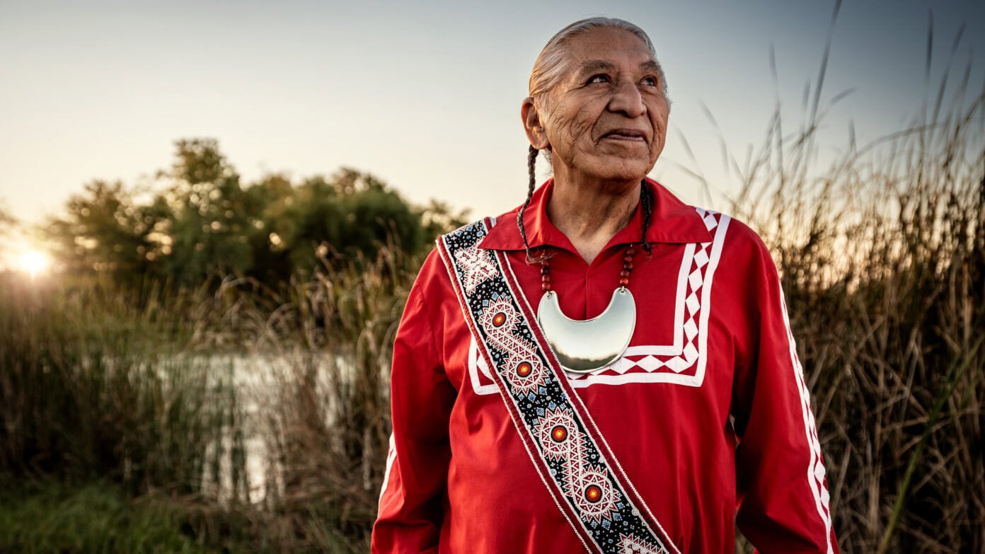 Wilson Roberts stands in front of reeds in traditional Choctaw clothing and jewelry