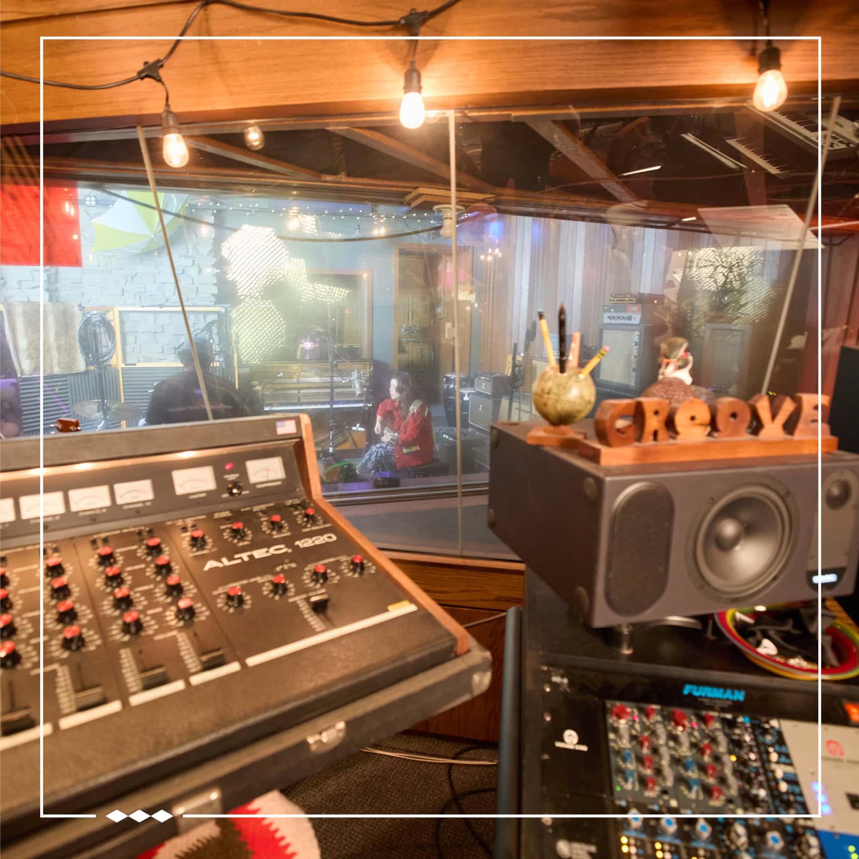 Samantha is seen recording guitar and vocals through the glass window of a music studio. A few mixing consoles and a speaker can be seen. The photo is inlaid with a white border with three diamonds in the bottom left corner.