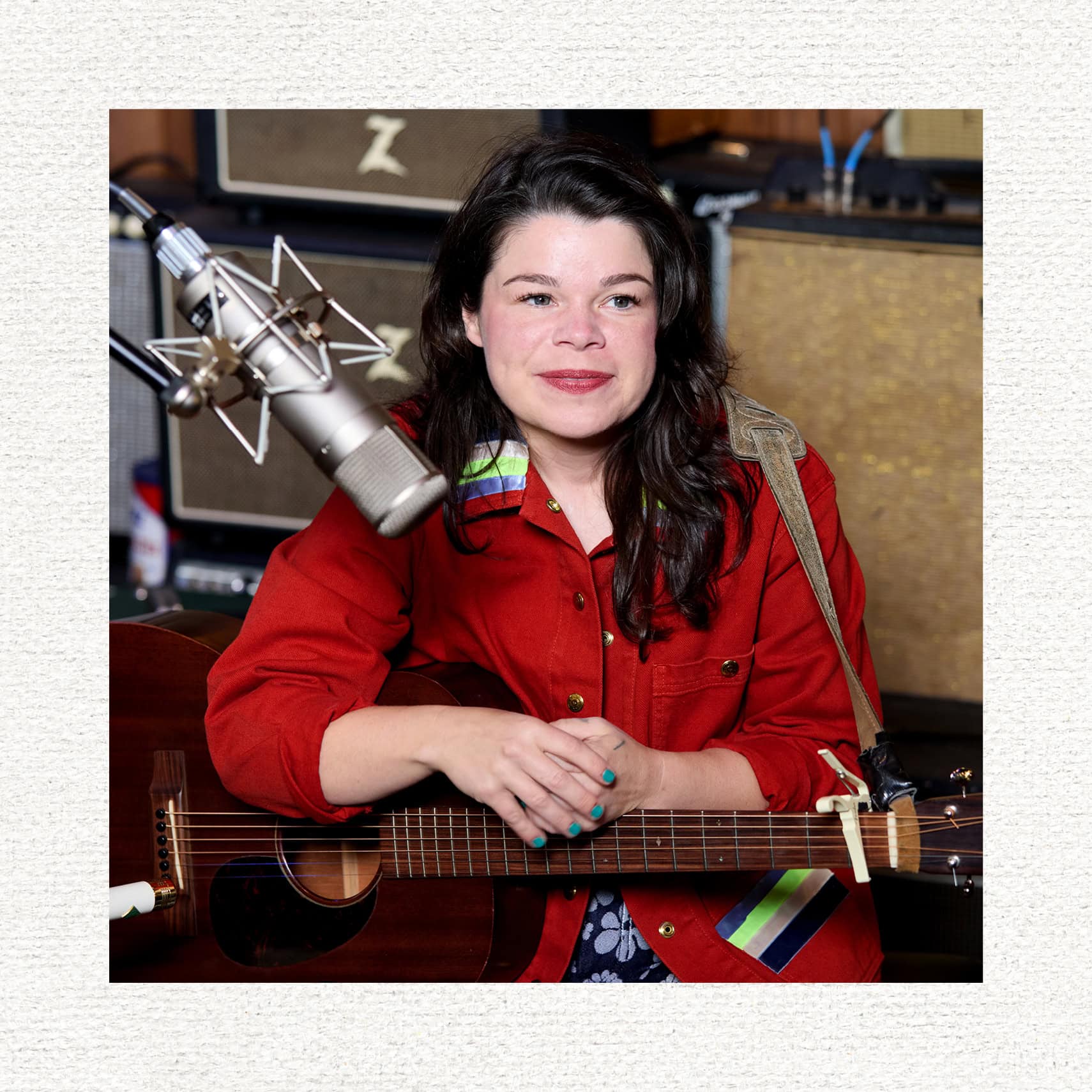 Samantha sits in front of a microphone, smiling and holding her acoustic guitar