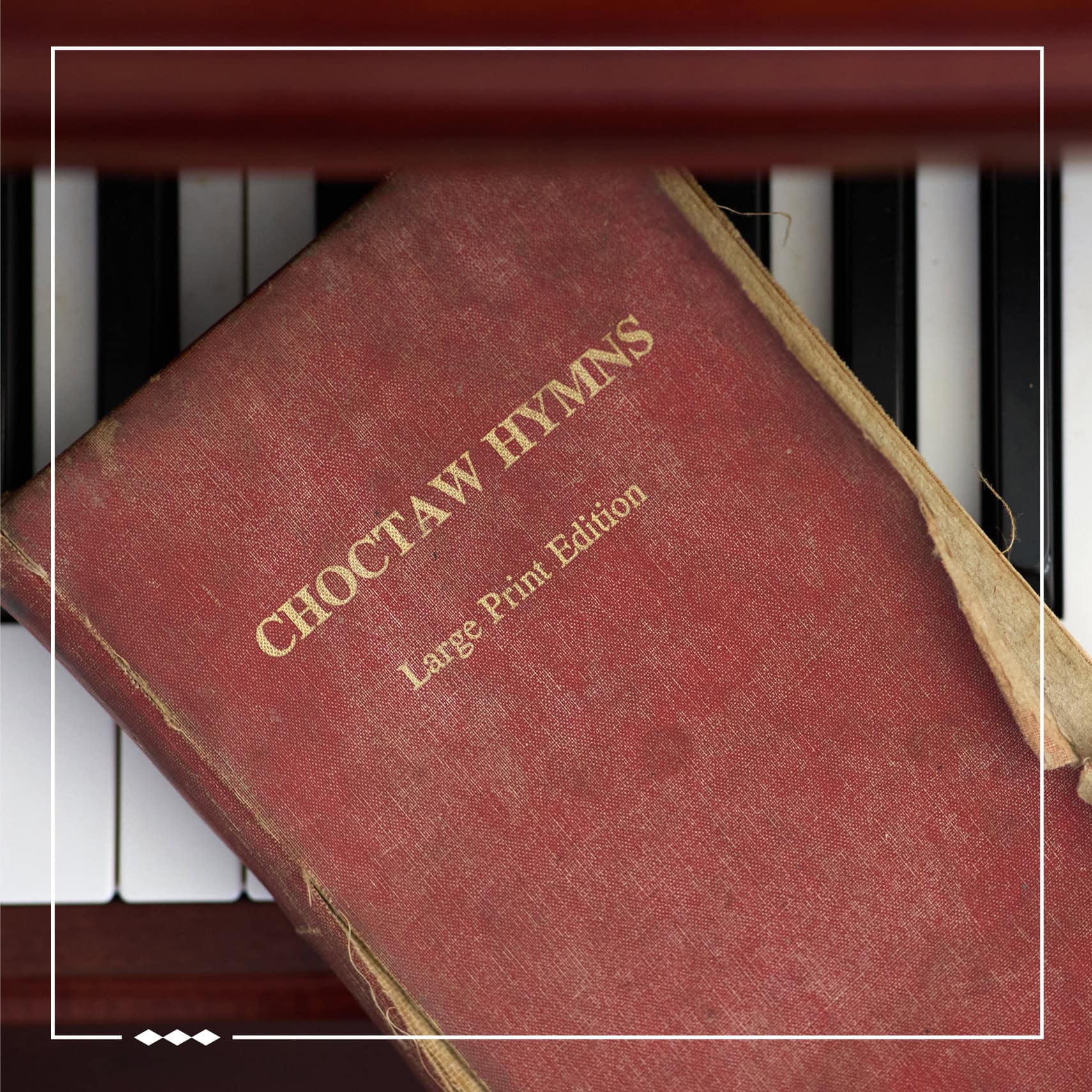 A Choctaw Hymnal sits on top of a piano. The photo is inlaid with a white border with three diamonds in the bottom left corner