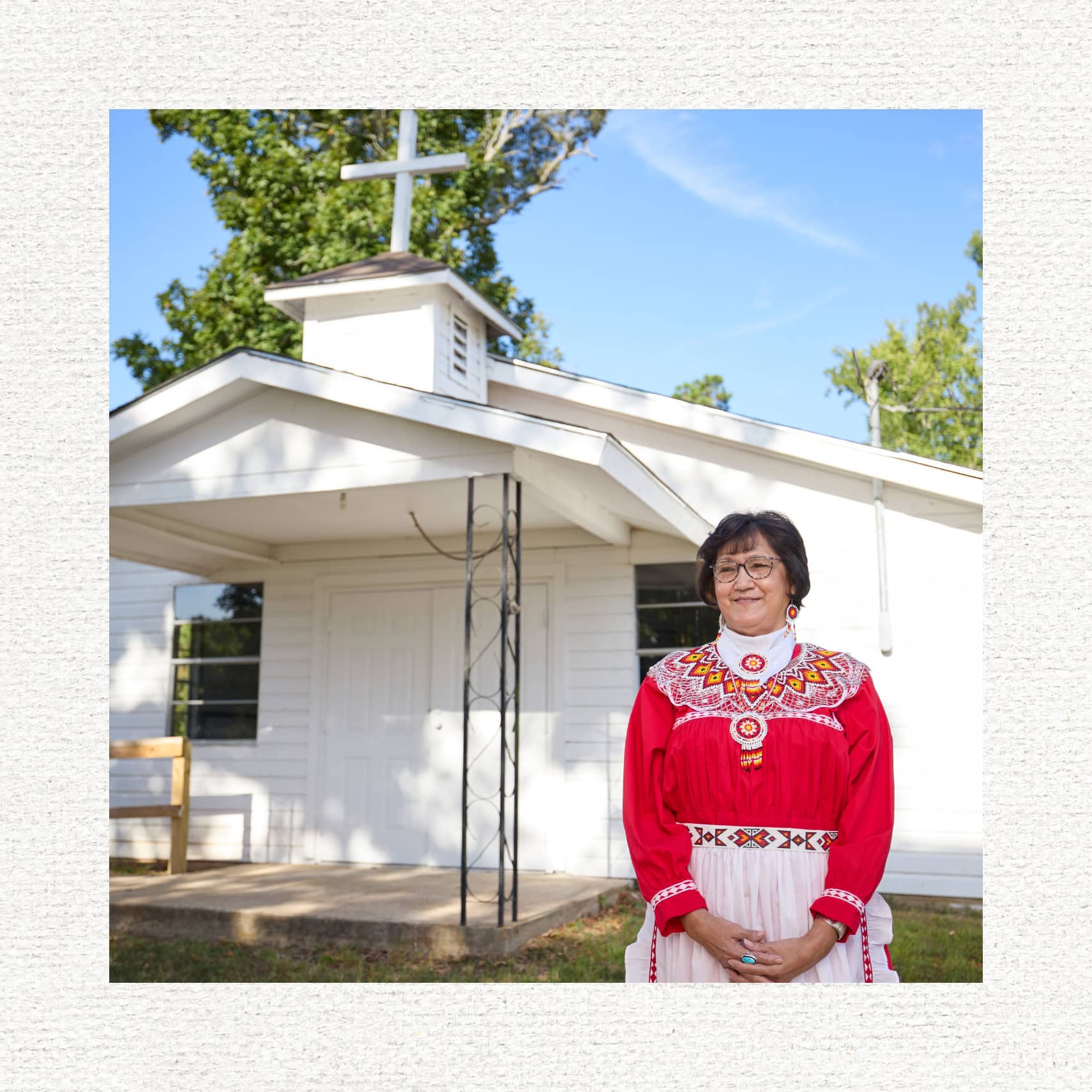 Terri Billy smiles in front of a church's entrance. She is wearing a red, traditional Choctaw dress with beaded earrings and necklace.